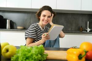 retrato de joven sonriente mujer en cocina, participación computadora portátil, haciendo notas para receta, escritura tienda de comestibles lista, Cocinando ensalada, sentado cerca vegetales y el cortar tablero foto