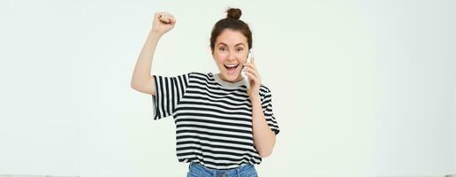 Portrait of enthusiastic young woman, talks on mobile phone, answers phone call and chanting, celebrating, laughing and smiling with excited face expression, white background photo