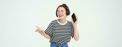 niña con teléfono inteligente baile, escuchando a música en inalámbrico auriculares, utilizando móvil teléfono transmisión aplicación a disfrutar favorito canciones, aislado terminado blanco antecedentes foto