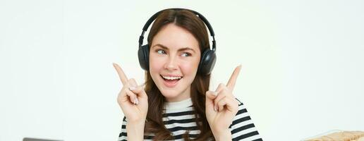 Dancing girl in wireless headphones pointing at empty space for music store promo text, showing advertisement, isolated over white background photo