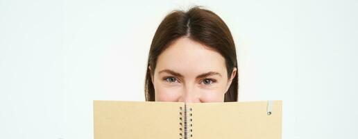Portrait of cute young woman hides her face behind planner, holds notebook against her face and smiles, isolated over white background photo