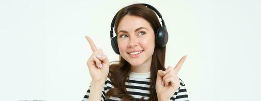 Close up portrait of beautiful girl dancing, listening music in wireless headphones and smiling, pointing sideways, showing two sides, advertisement or banner photo