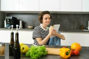 Portrait of beautiful modern woman, girl cooking with vegetables, holding notebook, writing down recipe, making notes, dinner plans, creating meal list for week, sitting in kitchen photo