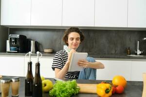 retrato de joven sonriente mujer Cocinando con verduras, escritura abajo ingredientes, mirando a su computadora portátil, leyendo receta, sentado en cocina foto