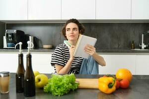 retrato de joven sonriente mujer en cocina, participación computadora portátil, haciendo notas para receta, escritura tienda de comestibles lista, Cocinando ensalada, sentado cerca vegetales y el cortar tablero foto