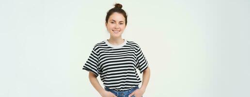 Image of charismatic young woman in striped t-shirt and jeans, looking confident and happy, smiling at camera, candid emotions, white background photo