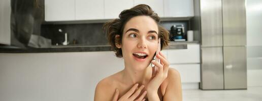 Close up portrait of young woman talking on mobile phone, looking surprised, hears amazing news, sitting near kitchen at home photo