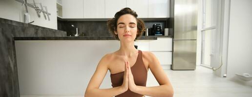 Portrait of female athlete, girl practice yoga, meditating in lotus pose, using mindfulness technique, workout at home on kitchen floor photo