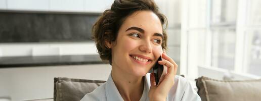 cerca arriba retrato de sonriente hermosa mujer, hablando en móvil teléfono, chats en teléfono inteligente, llamadas alguien, se sienta a hogar foto