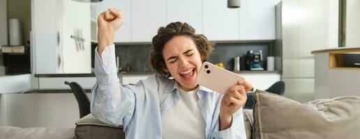 Image of girl celebrating, holding smartphone and cheering from excitement, winning, receive great news on mobile phone photo