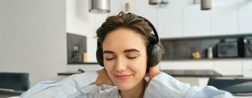 Close up portrait of girl with headphones, enjoys listen to music, close eyes and smiles form joy and delight photo