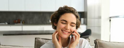 Close up portrait of happy brunette girl, talks on mobile phone, chats on smartphone, calls someone while spending time at home on sofa photo