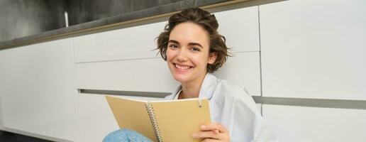 Portrait of happy young woman writes in her diary and smiles, makes notes in notebook, holds pen, sits on floor photo