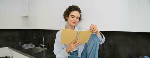 retrato de joven moderno mujer sentado en cocina, voltear páginas, leyendo diario libro, sonriente felizmente foto