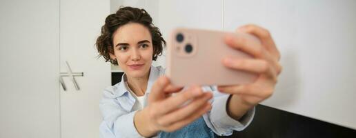 Stylish young girl blogger, takes selfie in her kitchen, poses for photo on mobile phone, using camera app for a video with filters