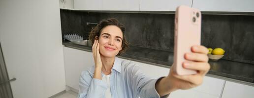 Image of stylish young woman blogger, recording video of herself, taking pictures on smartphone, making selfie at home photo