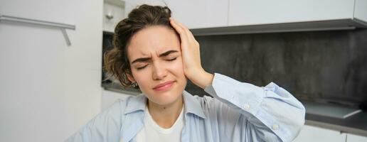 Close up portrait of brunette woman, touches her head, feels dizzy, has migraine or headache, grimaces from pain photo