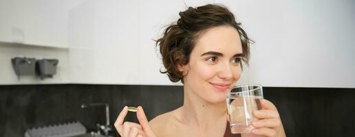 Portrait of sportswoman drinking water, taking vitamins, dietry supplements for healthy skin, having omega-3 pill, standing in her kitchen in workout clothing photo