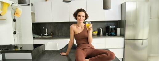 hermosa sonriente aptitud chica, vistiendo ropa activa, sentado en cocina encimera, Bebiendo vaso de naranja jugo y mirando contento foto