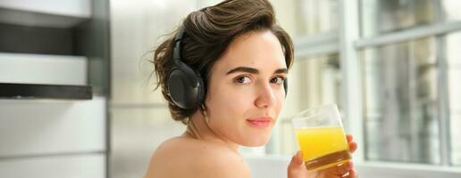 Sport and healthy lifestyle. Portrait of happy sporty girl, fitness woman in headphones, rewinds at home, drinks orange juice with happy smile after workout photo