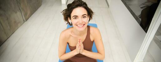 Relaxed brunette woman sitting in lotus pose on yoga mat, practice meditation, stretching exercises at home, workout on floor in leggings and top, sport and recreation concept photo