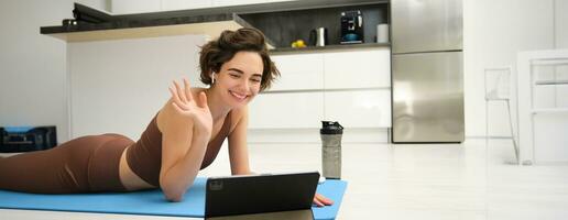 Smiling brunette woman in sportswear, waves hand, says hi at tablet, records video sports blog, fitness training session online, instructor shows workout elements, uses rubber mat at home in kitchen photo