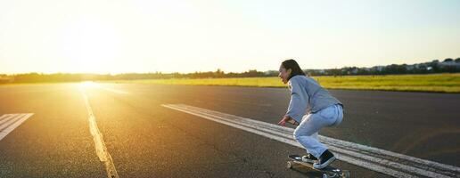 lado ver de hermosa asiático niña en patineta, montando su crucero hacia el Dom en un vacío la carretera. contento joven patinador disfrutando soleado día en su patinar foto