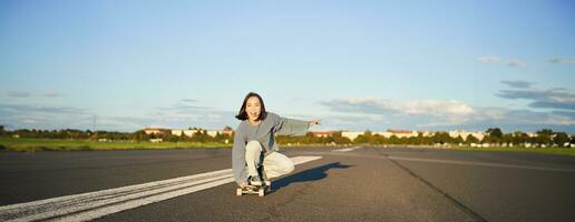 despreocupado patinador niña en su patineta, montando longboard en un vacío camino, participación manos oblicuo y riendo foto