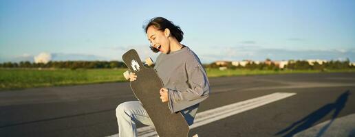 Beautiful asian teen girl playing with her longboard, holding skateboard as if playing guitar, standing on road on sunny day photo