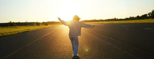 Teen girl feeling happy on longboard. Happy young skater riding her skateboard with hands spread sideways, feeling freedom, going towards the sun photo