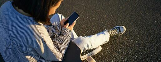 Close up of skater teen girl sits on skateboard, types message on smartphone, looks at mobile phone screen photo