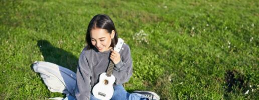 sonriente asiático niña aprende cómo a jugar ukelele en computadora portátil, vídeo charla con música maestro, sentado con instrumento en parque en césped foto