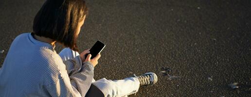 Vertical shot of asian girl typing, sending message or using mobile phone app, sitting on skateboard on road photo