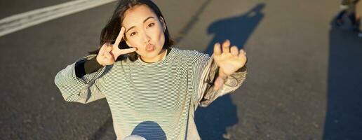 Selfie of asian girl sitting on skateboard, taking photo on smartphone, smiling and showing peace v-sign