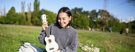 Vertical shot of happy asian girl plays instrument, shows her ukulele at laptop camera, video chats about music, teaches how to play, sits in park outdoors photo