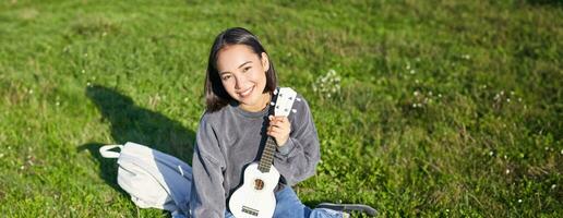 sonriente asiático niña aprende cómo a jugar ukelele en computadora portátil, vídeo charla con música maestro, sentado con instrumento en parque en césped foto