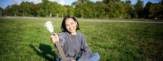 Music and instruments. Smiling asian girl shows her white ukulele, sits in park and plays small guitar photo