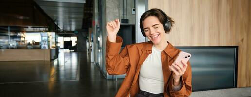 Close up portrait of happy employee, young office manager sitting on floor in break room, resting in co-working space, wearing wireless headphones, holding smartphone and dancing photo