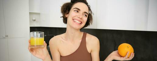 sano estilo de vida y deporte. hermosa sonriente mujer, Bebiendo Fresco naranja jugo y participación Fruta en su mano, disfrutando su vitamina bebida después rutina de ejercicio a hogar, en pie en cocina foto