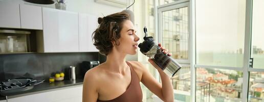 hembra atleta en su cocina, en pie con agua botella, Bebiendo después rutina de ejercicio yoga capacitación. ropa de deporte en Deportes vestir corsé hidratado después gimnasio ejercicio foto