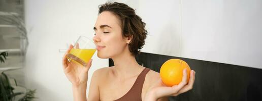 Image of sportswoman, fitness girl holding glass of juice and an orange, smiling, drinking vitamin beverage after workout, standing in her kitchen at home. Healthy lifestyle and sport concept photo