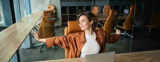 Portrait of woman resting in office, stretching arms after sitting and working on laptop photo