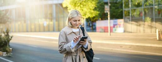 Image of young woman on street, carries backpack, looks at her smartphone, using carsharing application, orders taxi, sends a message on mobile app photo