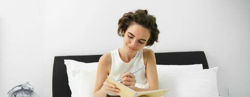 Image of mindful young woman sitting in bed, waking up and writing in journal, making notes, sharing her thoughts in diary photo