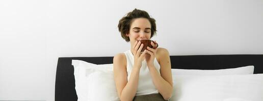 hermosa joven mujer despertar arriba y Bebiendo café en su cama, sonriente desde satisfacción. foto