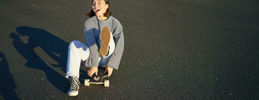 Portrait of beautiful asian girl skating, sitting on her skateboard and smiling. Cute teenager with longboard, skating on road photo