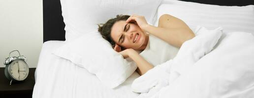 Portrait of woman with headache, lying in bed and suffering from migraine, touching head and grimacing from discomfort photo