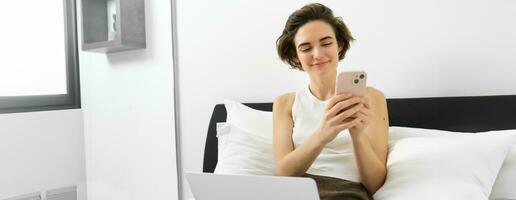 Image of stylish modern woman, sitting on bed, resting at home with laptop and smartphone, sending message, reading notification on mobile phone photo