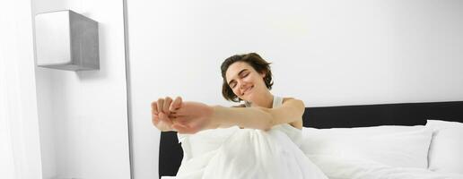 Portrait of smiling young woman wakes up in her bed, sitting under white linen sheets with closed eyes and stretching her hands with satisfaction photo