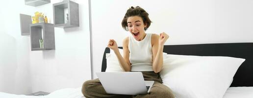 Portrait of woman sitting on bed, looking at laptop with excited, amazed face, celebrating, making fist pump, triumphing after winning on computer photo